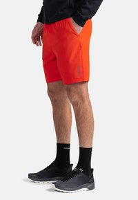 Shorts sportivi di un brillante colore arancione con una superficie liscia, dotati di tasche laterali e di un discreto logo del marchio. Indossati con scarpe da ginnastica nere e calze.