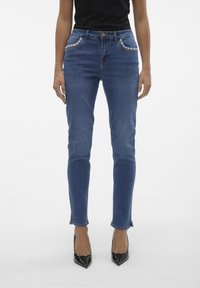 Jeans ajustados de mezclilla azul con cintura de media altura, que presentan detalles bordados en los bolsillos y un ligero deshilachado en el dobladillo.