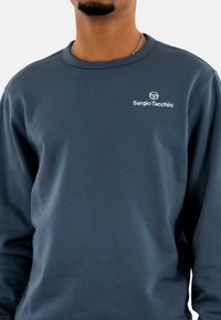 Sudadera azul oscura hecha de tela suave. Tiene un cuello redondo, mangas largas y un logo blanco en el pecho.