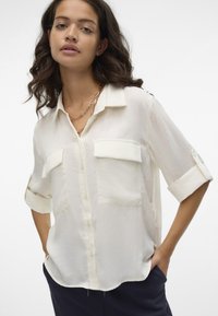 Svetlý slonovinový blouse s krátkymi rukávmi, zapínanie na gombíky vpredu, dve náprsné vrecká a mierne priesvitná textúra. Jednoduchý golier, voľný strih.