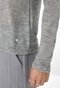 Grau gestrickter Pullover mit Paillettenakzenten, gerippten Bündchen und Saum, präsentiert mit grauen gestreiften Hosen. Nahaufnahme von Ärmeldetail und Hand.