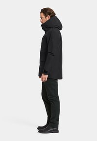 Svart vattentät jacka med hög krage och stor huva, med sidofickor och justerbara ärmslut. Bärs med mörka byxor och stövlar.