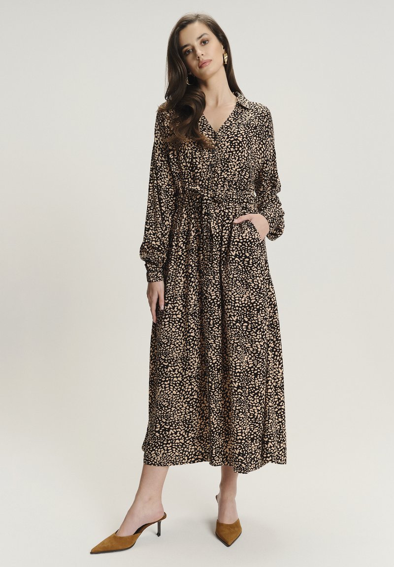 Lungo vestito nero con un sottile motivo a macchie crema e marrone, colletto con bottoni, vita elasticizzata e maniche lunghe, abbinato a tacchi beige.