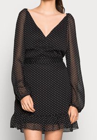 Černé šaty s bílými puntíky, ve výstřihu do V, s dlouhými průhlednými volánovými rukávy a volánkovým lemem. Vyrobeno z lehké, splývavé tkaniny.