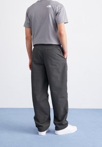 Graues T-Shirt mit kleinem Logo auf dem Rücken, kombiniert mit lockeren, dunkelgrauen Hosen. Weiße Sportschuhe runden das Outfit ab.