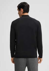 Maglione nero lavorato a maglia con colletto alto, maniche raglan e polsini a coste. Il retro è liscio, indossato da un modello con pantaloni grigi.
