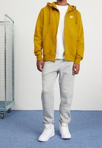 Sudadera amarilla con cremallera y cordones, logo blanco, combinada con pantalones de chándal gris claro y zapatillas blancas de caña alta sobre una alfombra azul.