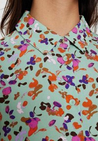 Camicia button-up in tessuto verde menta con un motivo floreale multicolore in viola, arancione, rosa e marrone. Dettaglio del colletto visibile.