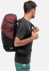 Homme ajustant les sangles d'un sac à dos de randonnée bordeaux et noir, portant un t-shirt noir et un short bleu-vert, tourné vers la droite avec un fond neutre.