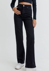 Jean évasé en denim noir taille haute avec fermeture à boutons à l'avant et poches, associé à des baskets blanches sur un fond neutre.