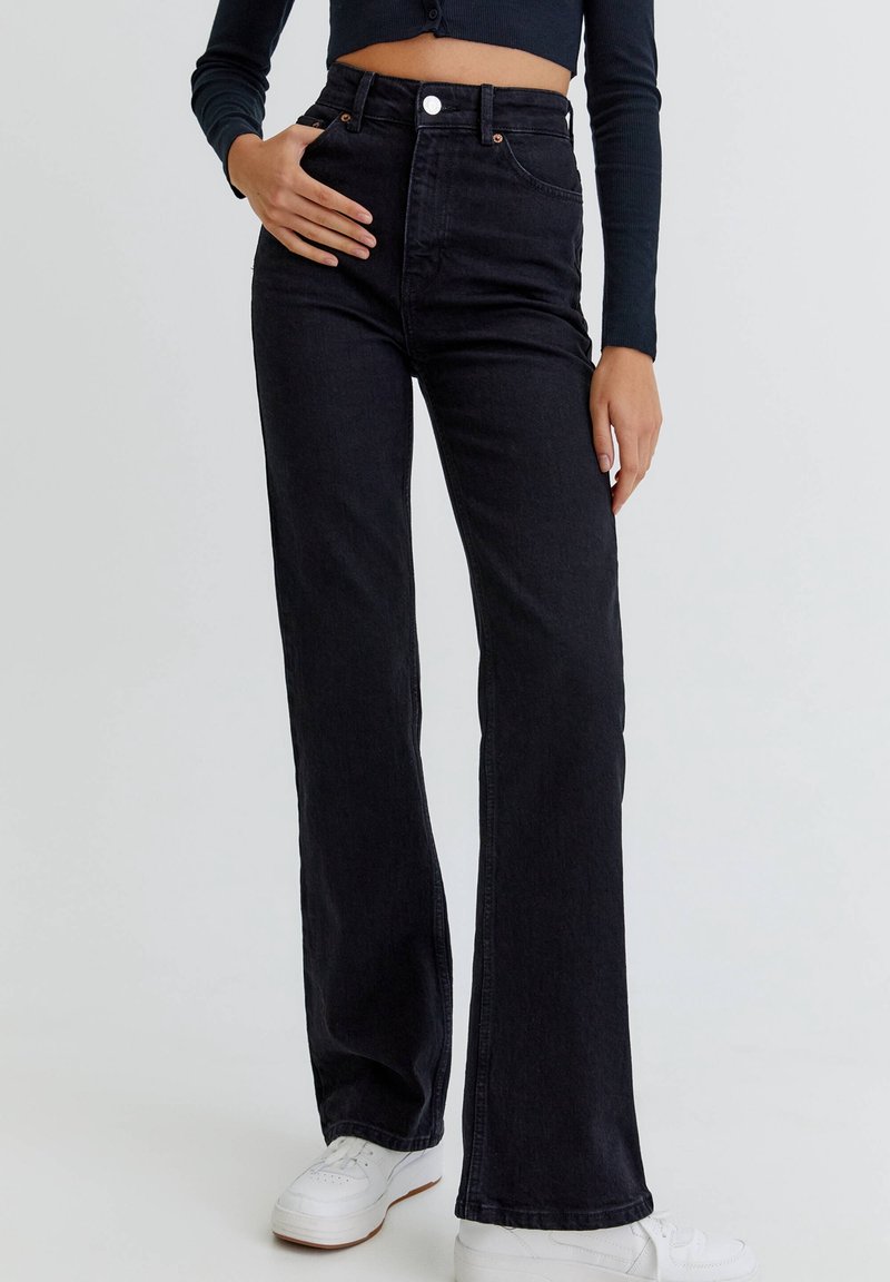 Jean évasé en denim noir taille haute avec fermeture à boutons à l'avant et poches, associé à des baskets blanches sur un fond neutre.