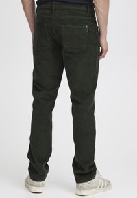 Donkergroene ribfluweelbroek met een ribbelstructuur, voorzien van twee achterzakken en een ritsdetail. De pasvorm is taps toelopend en casual.