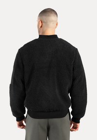 Hombre con cabello corto vistiendo una chaqueta de forro polar texturizado negro y pantalones grises, de espaldas frente a un fondo claro y liso.