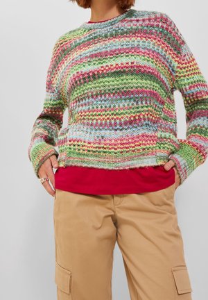 Personne portant un pull en maille rayé multicolore par-dessus une chemise rouge et un pantalon cargo beige, mains dans les poches, torse visible.
