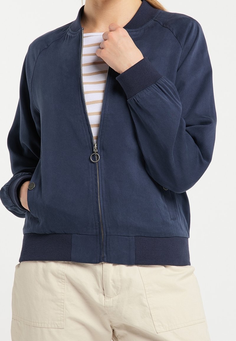 Personne portant une veste bomber zippée bleu marine sur une chemise rayée blanche et beige avec un pantalon beige, main dans la poche et tenant le col de la veste.