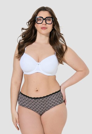 Mujer con cabello largo y castaño, que lleva grandes gafas negras, un sujetador blanco y ropa interior estampada en negro y beige, de pie frente a un fondo liso.