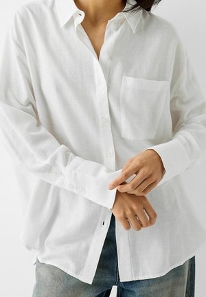 Persoon die een losse witte blouse met borstzak draagt en met de handen de manchetten verstelt, gecombineerd met een blauwe spijkerbroek.