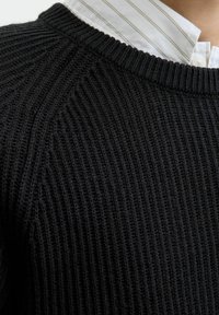 Zwarte gebreide trui met ribbelstructuur, met een ronde halslijn. Een witte gekraagde blouse met dunne strepen is zichtbaar eronder.