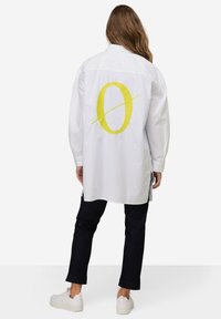 Camicia bianca oversize con un grande design giallo a forma di 'O' sul retro. Presenta maniche lunghe e orlo dritto. Indossata con jeans scuri e scarpe bianche.
