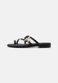Svart platt sandal med vävd struktur, smala remmar och en dekorativ guldaccent. Enkel, öppen design med låg klack och minimalistisk stil.