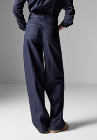 Tummat farkut leveillä lahkeilla, joissa on kaksi takataskua, suora leikkaus ja sileä pinta. Yhdistettynä vaaleanvärisiin jalkineisiin.