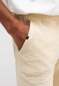 Beige strukturierten Hosen mit einem gewebten Muster, ausgestattet mit einem elastischen Taillenbund und seitlichen Taschen. An der Taschenkante ist ein kleines schwarzes Etikett sichtbar.