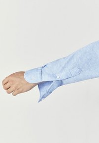 Vaaleansininen pitkähihainen paita, jossa on tekstuuri. Hihassa on nappilisä ja näkyvä laskostus.