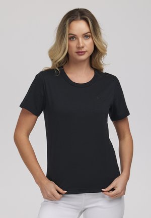 Kobieta o blond włosach, ubrana w jednolity czarny t-shirt z krótkim rękawem i białe spodnie, stojąca na jasnoszarym tle.