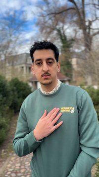 Ung mand udendørs iført grøn "Bermuda Shore" sweatshirt, hånd på brystet, med slørede træer og bygning i baggrunden.