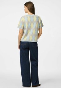 Suéter de manga corta, color azul claro, con un patrón de diamantes en amarillo y gris. Presenta un cuello acanalado y un corte relajado, combinado con jeans oscuros.