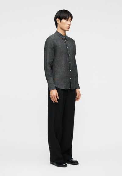 Νεαρός άνδρας όρθιος, φορώντας σκουρόγκρι πουκάμισο με κουμπιά, άνετο μαύρο παντελόνι και μαύρα παπούτσια, σε απλό ανοιχτό φόντο.