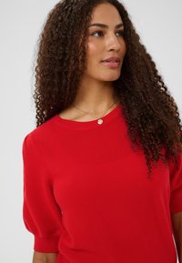 Pull rouge en maille avec manches courtes bouffantes, encolure côtelée et tissu texturé. Collier pendentif doré apportant un détail subtil.