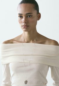 Beżowa bluzka z odkrytymi ramionami wykonana jest z miękkiego materiału z teksturowanymi fałdami, uzupełniona srebrnym wisiorkiem na delikatnym łańcuszku.