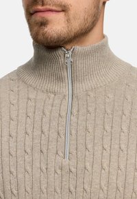 Beige kabelstrikket genser med h øy ribbestrikket krage og en sølvglidelås ved halsen, som viser en myk tekstur og detaljert mønster.