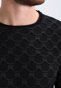 Pull en tricot noir avec un motif texturé de cercles superposés, col rond et poignets côtelés. Gros plan sur les détails du tissu.