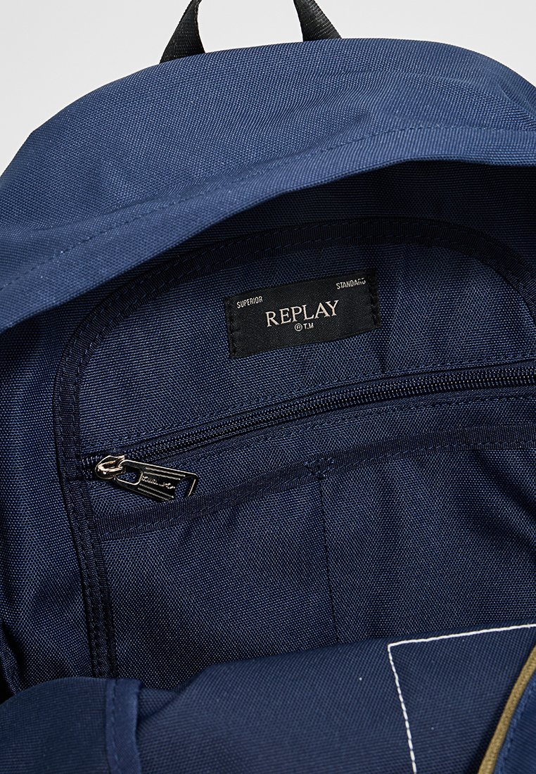 Interno di uno zaino blu navy, dotato di una tasca con zip e un'etichetta con la scritta "REPLAY." Il materiale è una stoffa texturizzata con cuciture rinforzate.