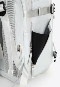 Sac à dos gris clair avec des accents en maille, plusieurs zippers, une poche latérale noire, et des sangles argentées pour l'attache et l'ajustement.