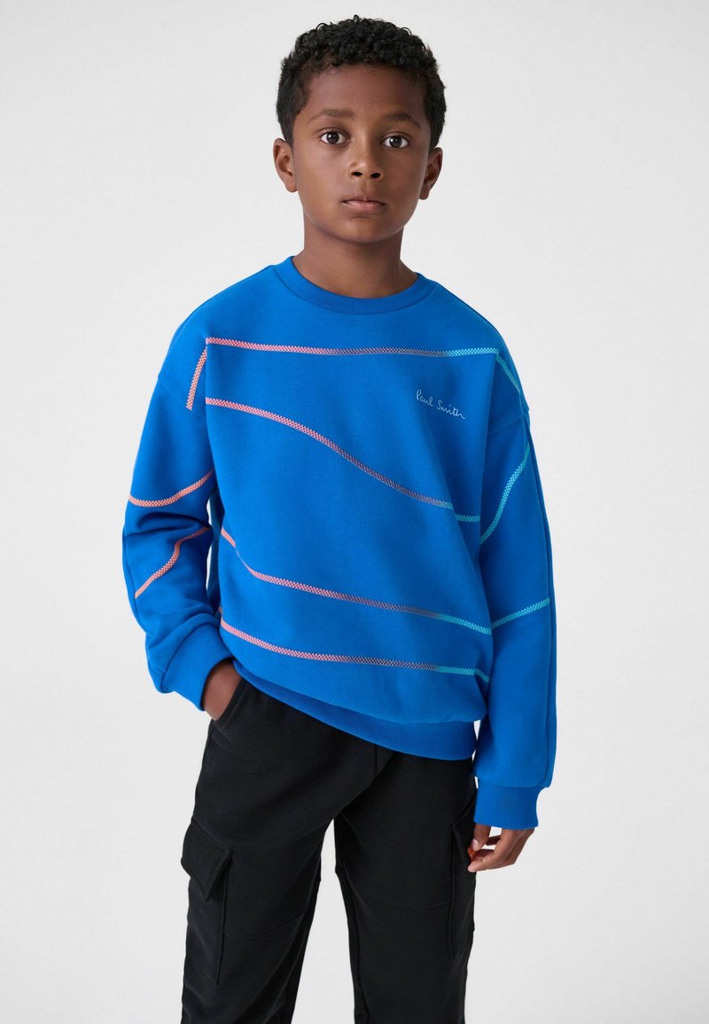 Modrá mikina so horizontálnymi vlnovými vzormi v ružovej a tyrkysovej farbe, okrúhly výstrih a dlhé rukávy, kombinovaná s čiernymi cargo nohavicami.