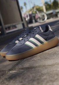 Par de zapatillas Adidas azul oscuro con rayas blancas y verdes y suelas de goma translúcidas sobre un pavimento de piedra al aire libre.