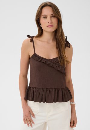 Femme portant un haut marron sans manches avec des volants et des bretelles nouées aux épaules, associé à un pantalon blanc sur un fond uni.