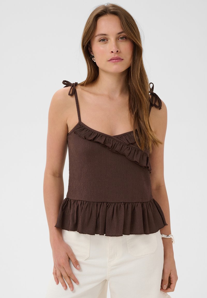 Femme portant un haut marron sans manches avec des volants et des bretelles nouées aux épaules, associé à un pantalon blanc sur un fond uni.