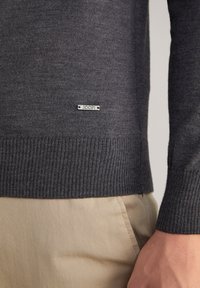 Dunkelgrauer Strickpullover mit geripptem Saum und Bündchen; verfügt über ein silbernes Metallschild mit dem "JOOP!"-Logo an der Unterseite.