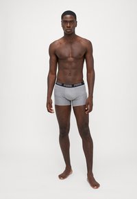 Szürke boxeralsó fekete, rugalmas derékpánttal, amelyen a "PUMA" logó látható, sima textúrával és minimalista dizájnnal. A modell mezítláb áll.