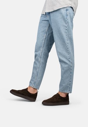 Scarpe slip-on in suede marrone con finitura texturizzata, abbinate a jeans in denim a lavaggio chiaro e vestibilità rilassata. Suola in gomma nera solida.