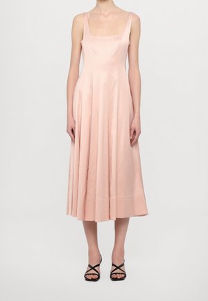 Kobieta ubrana w różową sukienkę midi bez rękawów z kwadratowym dekoltem oraz czarne sandały na wysokim obcasie z paskami, stojąca na tle jednolitego tła.