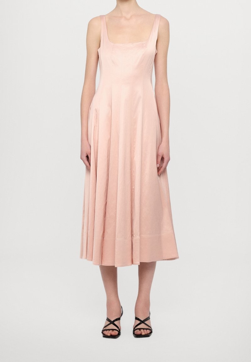 Femme portant une robe midi rose poudré sans manches avec un décolleté carré et des sandales à talons hauts noires à lanières, debout devant un fond uni.