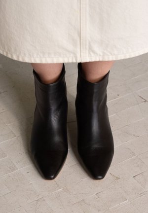 Stivaletti alla caviglia in pelle nera con punta appuntita e texture liscia, visti dall'alto su una superficie chiara e texturizzata.