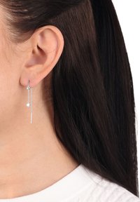 Silverhängande stjärna och kedjeörhängen bärs i vänster öra, med mörkt hår och en vit ribbad topp synlig.