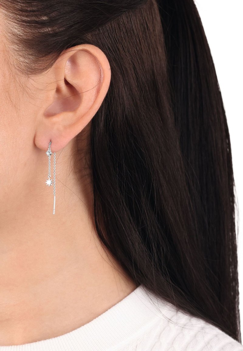 Silverhängande stjärna och kedjeörhängen bärs i vänster öra, med mörkt hår och en vit ribbad topp synlig.