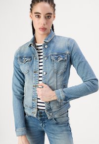 Veste en denim bleu au design classique, avec des fermetures à boutons argentés, deux poches poitrine et une texture légèrement délavée. Haut rayé visible en dessous.